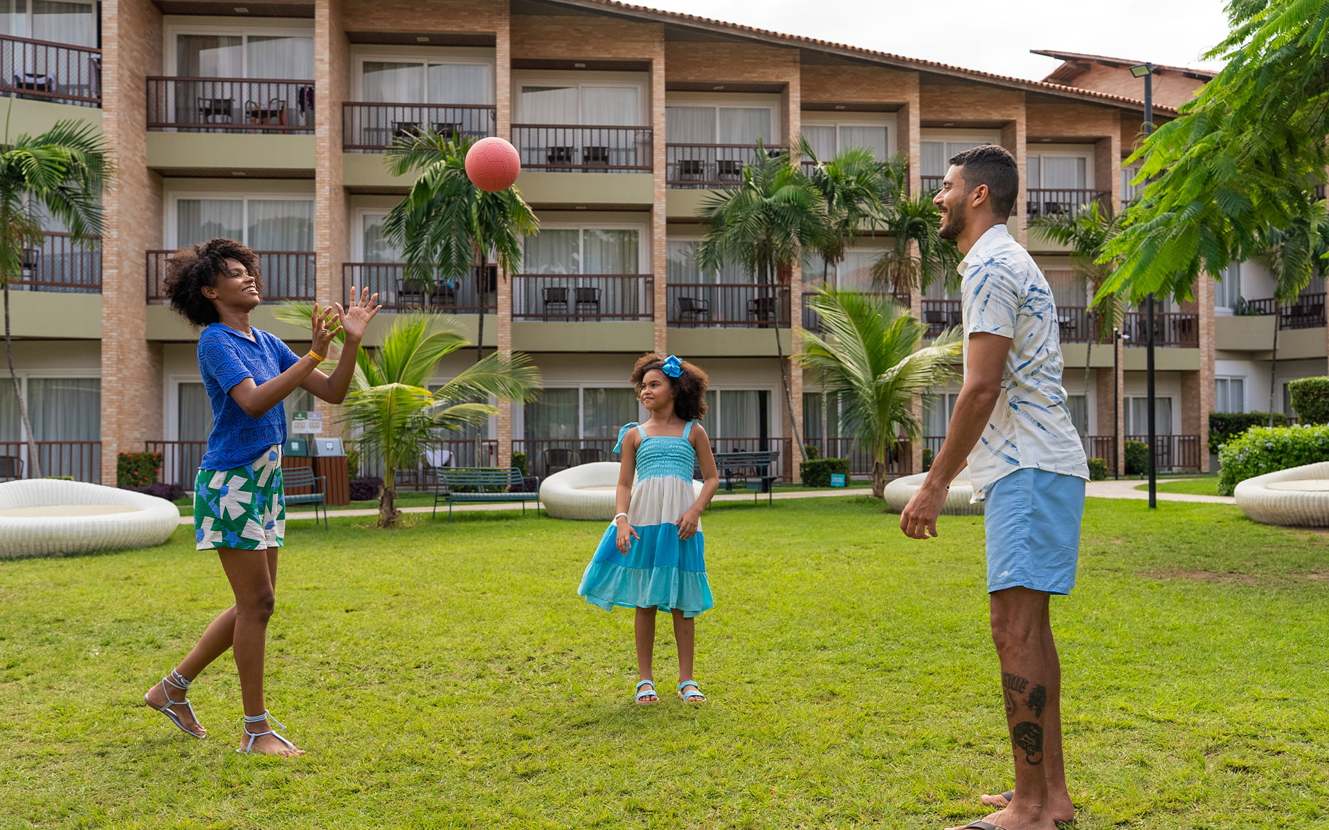 Atividades recreativas pra toda a família Família brincando de bola no gramado ao ar livre no Salinas Maceió, com toda estrutura dos apartamentos ao fundo.