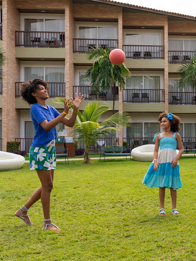 Atividades recreativas pra toda a família Família brincando de bola no gramado ao ar livre no Salinas Maceió, com toda estrutura dos apartamentos ao fundo.
