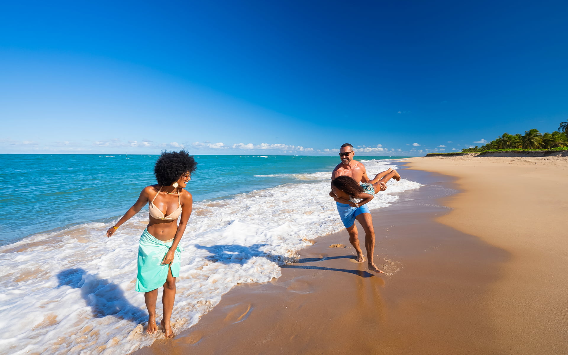 Momentos inesquecíveis na Praia do Gunga Família caminhando na Praia do Gunga com mãe a frente, e pai brincando de avião com a filha na faixa de areia com dia ensolarado.