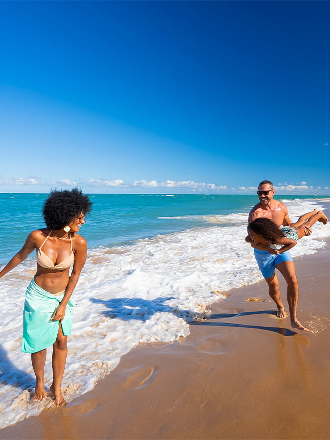 Momentos inesquecíveis na Praia do Gunga Família caminhando na Praia do Gunga com mãe a frente, e pai brincando de avião com a filha na faixa de areia com dia ensolarado.