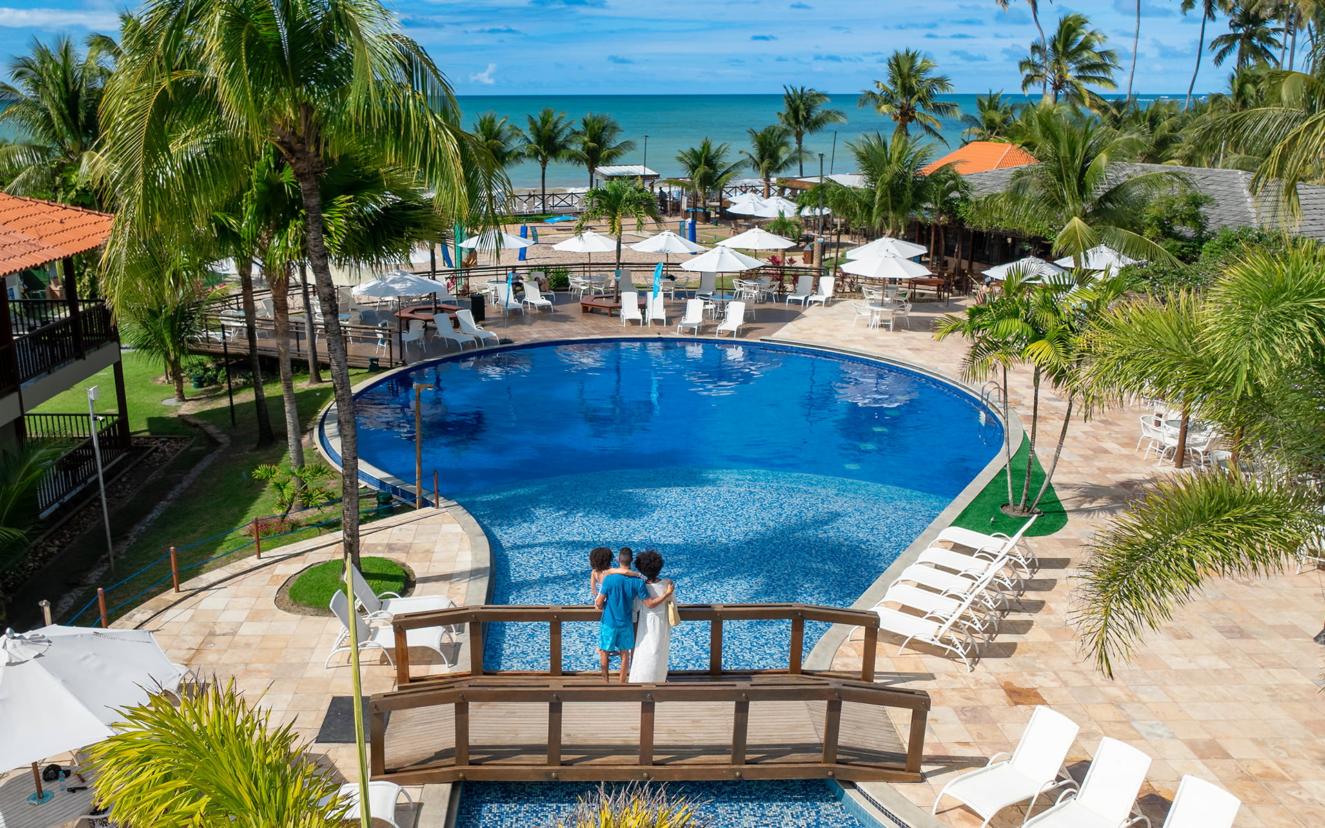 Piscinas do Salinas Maceió pra todas as idades Piscina redonda, com ponte de madeira passando por cima dela. Na ponta há uma família de pai, mãe e filha observando o horizonte do resort e praia do Salinas Maceió cercada por coqueiros e áreas de lazer.