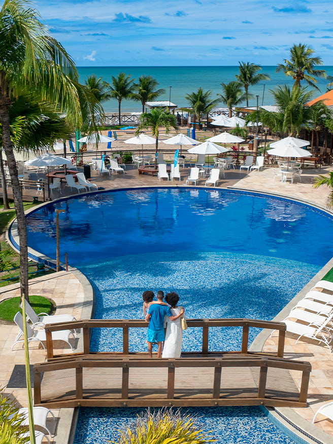 Piscinas do Salinas Maceió pra todas as idades Piscina redonda, com ponte de madeira passando por cima dela. Na ponta há uma família de pai, mãe e filha observando o horizonte do resort e praia do Salinas Maceió cercada por coqueiros e áreas de lazer.