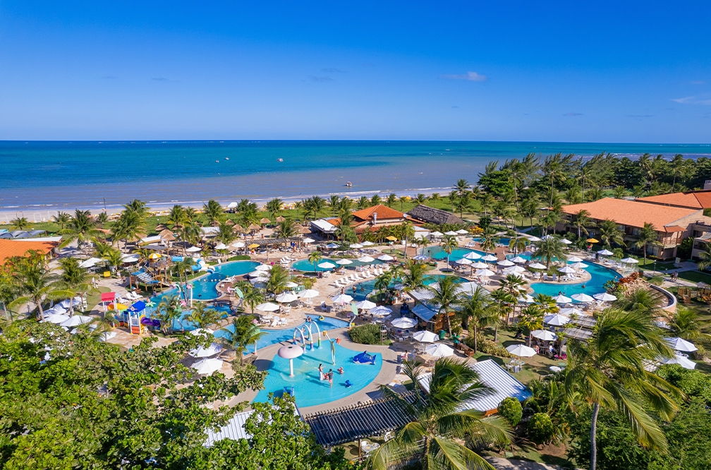 Estrutura completa com diversão pra todos Vista aérea do complexo de piscinas do Salinas Maceió, é possível ver brinquedos infantis, guarda-sois, e pessoas se divertindo. Com muitas árvores ao redor, e ao fundo a praia de Maragogi.