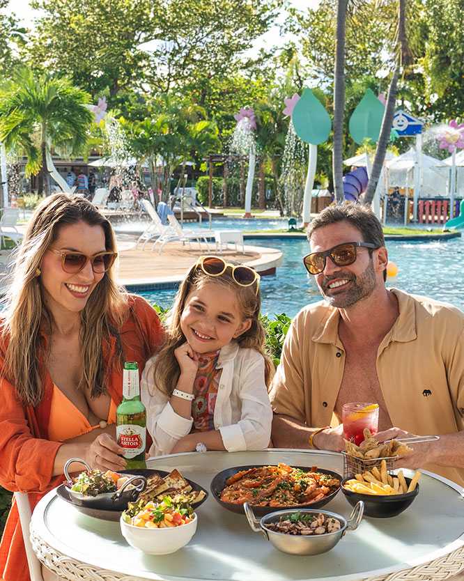 familia desfruta petiscos na piscina salinas maragogi Família sorridente com pai, mãe e filha fazendo um lanche em uma mesa branca ao lado da piscina com brinquedos aquáticos infantis do resort Salinas Aaragogi. Sobre a mesa, há porções de camarão, batata frita e bolinhos.