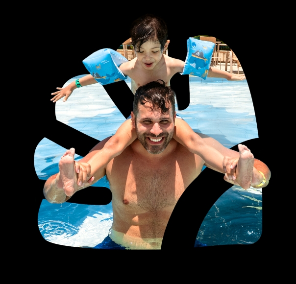 Filho sentado nas costas do pai, ambos estão sorrindo. O filho está com boias nos dois braços e com as pernas estiradas e o pai segurando elas. Eles estão dentro da piscina com o sol refletindo e no fundo é possível ver algumas cadeiras de madeira na beira da piscina.