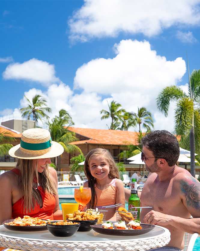 all inclusive salinas maragogi Família sorridente com pai, mãe e filha posando pra foto em uma mesa com diversos pratos de comida e bebidas, ao lado da área da piscina. Ao fundo, algumas espreguiçadeiras, guarda-sóis, coqueiros e a estrutura do Salinas Maragogi.
