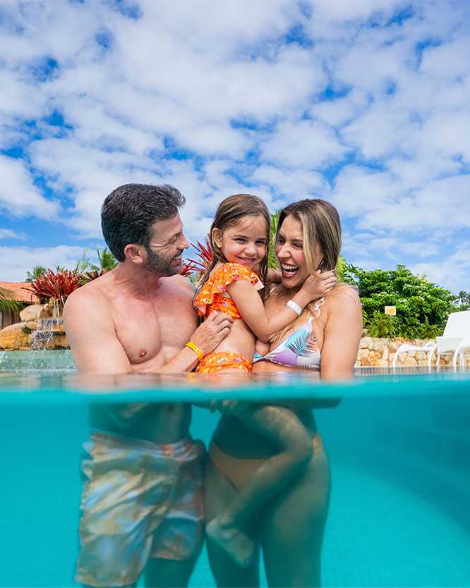 diversao em familia no salinas maragogi Família feliz, composta por pai, mãe e filha pequena, abraçados dentro da piscina azul do resort. A foto é tirada na altura da água, mostrando o céu azul com nuvens ao fundo, coqueiros e a arquitetura do Salinas Maragogi.
