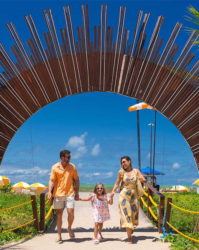 familia portal praia salinas maragogi Família com pai, mãe e filha pequena de mãos dadas, caminhando sorridentes em direção à câmera, passando por dentro de um grande arco de madeira que serve de portal pra a praia do resort, com coqueiros e o céu azul ao fundo.