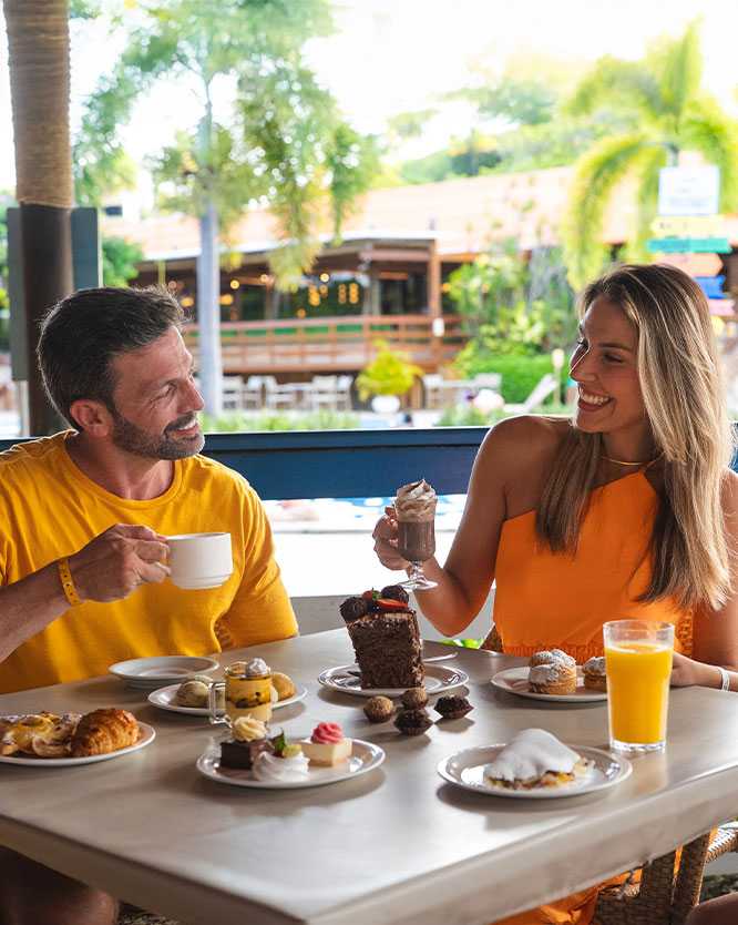 all inclusive para familia Família sorrindo e desfrutando de um lanche com muitos doces, salgados e bebidas em uma mesa. A mãe segura um copo com chantilly, o pai uma xícara e a filha ta na frente de um suco. A mesa está cheia de bolos, doces finos e salgados.