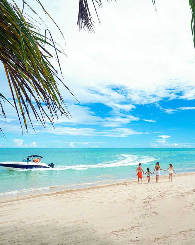 férias em família no salinas maragogi Família com dois adultos e duas crianças caminhando pela faixa de areia de uma praia paradisíaca, com o mar azul-turquesa ao lado. Uma lancha navega na água, e a cena é emoldurada por folhas de coqueiro em um dia de céu azul.