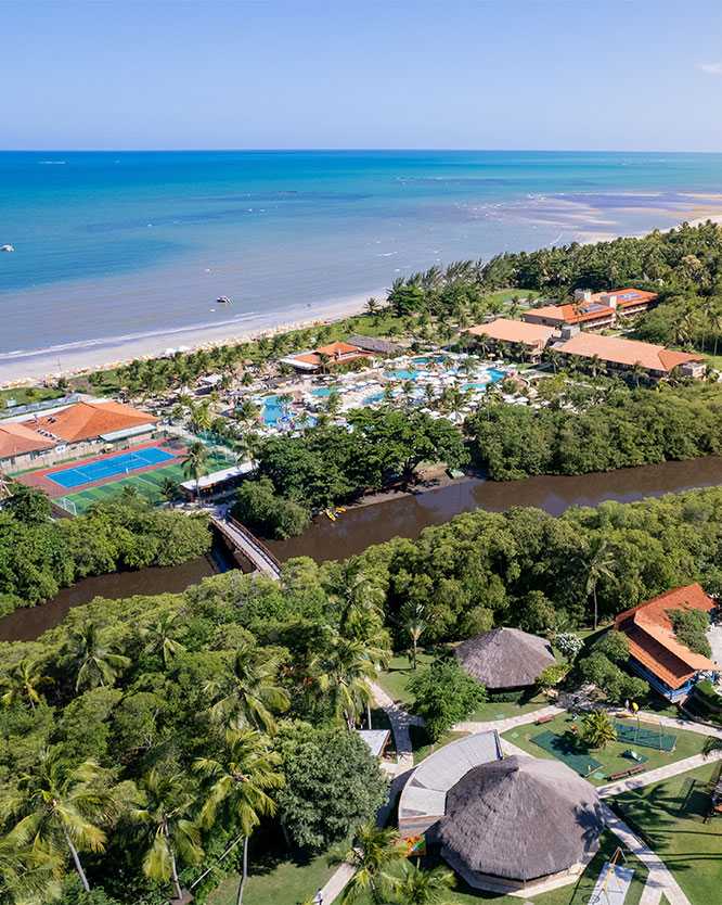 vista aerea salinas maragogi Vista de ângulo elevado do complexo do Salinas Maragogi, mostrando uma área de lazer infantil, quadras, piscinas maiores, estrutura do resort, com o rio cortando o resort com uma ponte ligando os dois lados, e o mar azul com a praia ao fundo, tudo cercado por coqueiros.