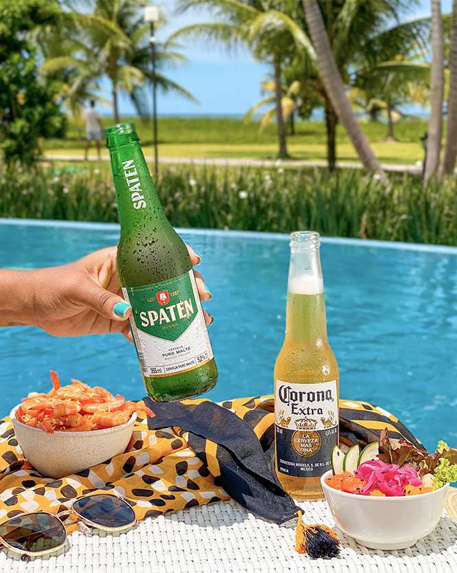 salinas maragogi all inclusive Mão segurando uma garrafa de cerveja Spaten. Sobre uma canga colorida na borda da piscina, estão uma garrafa de Corona Extra, óculos de sol e dois bowls de petiscos frescos. Ao fundo, coqueiros e o mar.